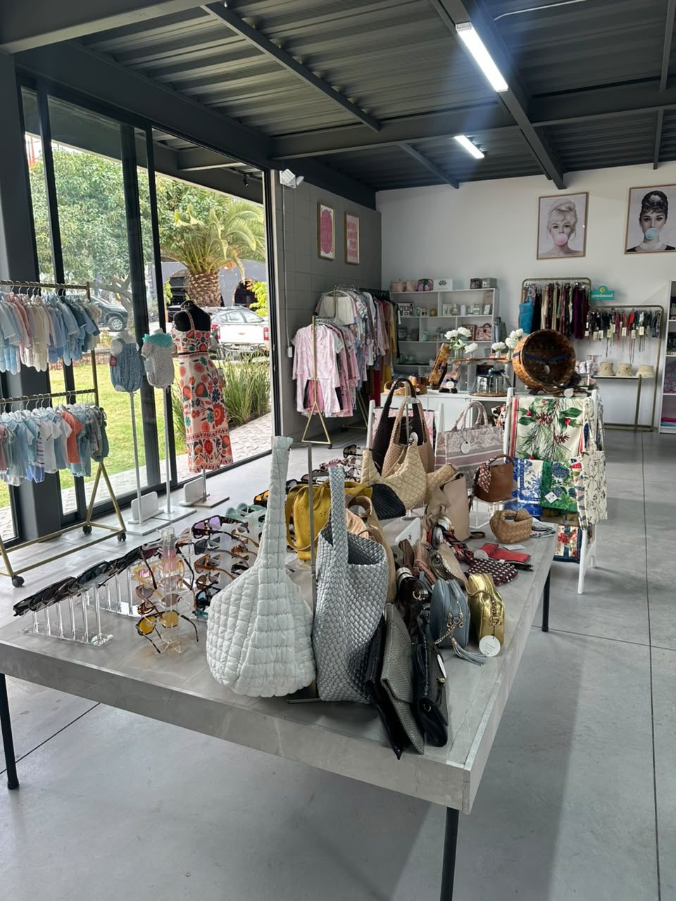 Mesa central con carteras, bolsos tejidos y gafas de sol; a la izquierda, perchas con ropa de bebé junto al ventanal